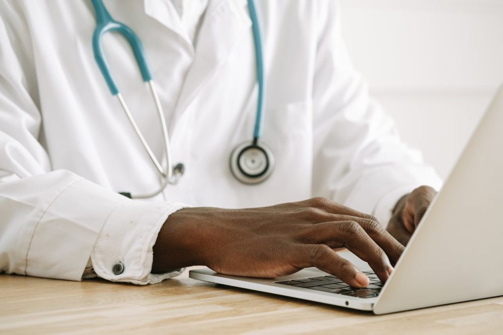 Close-up of a doctor using a laptop, wearing a stethoscope and white coat.